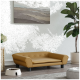 Ein stilvolles, hellbraunes Haustiersofa mit Knopfdetails steht auf einem Holzboden, neben einer weißen Futterschüssel, die mit Trockenfutter gefüllt ist, und einer Topfpflanze.