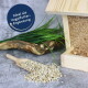 Holz-Vogelfutterspender mit herausfallenden Samen, umgeben von Gras; der Text lautet "Ideal als Vogelfutter-Ergänzung."