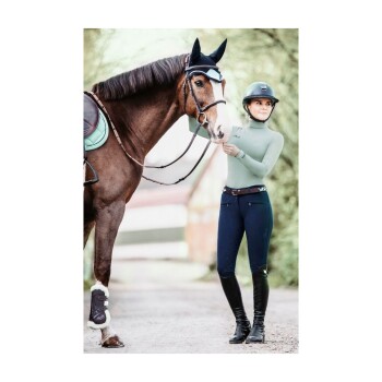 Ein Reiter in einem hellgrünen Hemd und dunkelblauen Hosen steht neben einem braunen Pferd mit Sattel und Beinschutz und hält dessen Zügel.