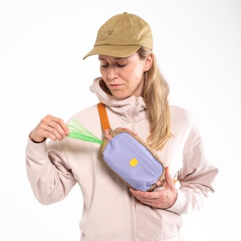 Eine Person mit einer beigen Mütze und einer pinken Jacke hält einen lila Hundekotbeutelspender mit einem grünen Beutel, der teilweise herausragt, getragen mit einem orangefarbenen Riemen.