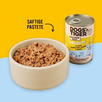 Eine beige Schüssel, gefüllt mit feuchtem Haustierfutter, steht neben einer Dose mit der Aufschrift "DOGS'n TIGER FREIGEIST," die Huhn mit Lachs und Karotten enthält.