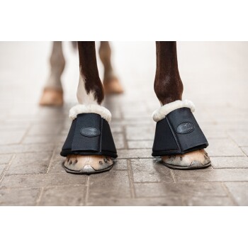 Die Vorderbeine eines Pferdes, die schwarze Hufschuhe mit weißem Fleecefutter tragen, stehen auf einer Ziegeloberfläche.