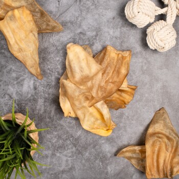 Ein Haufen getrockneter Tierohren-Leckerlis für Haustiere auf einer grauen Oberfläche, begleitet von einem kleinen geflochtenen Korb mit grünem Gras und einem weißen Seilspielzeug.