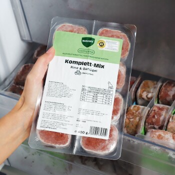 Hand hält eine gefrorene BARFER'S Komplett‑Mix Rind & Geflügel Katzenfutter‑Packung (400 g) im Gefrierschrank, mit sichtbaren Fleischstücken.