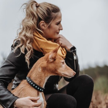Eine Frau in einer schwarzen Lederjacke und einem gelben Schal sitzt im Freien und hält sanft einen braunen Hund mit einem blauen, mit Nieten besetzten Halsband.