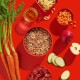 Ein roter Hintergrund zeigt eine Schüssel mit Tierfutter mit Fleisch, Gemüse und Pasta, umgeben von Karotten, einem Apfel und Gurkenscheiben.