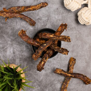 Eine schwarze Schüssel gefüllt mit Hundekauartikeln aus getrockneten Fleischstangen, umgeben von verstreuten Kauartikeln und einer kleinen grünen Pflanze.