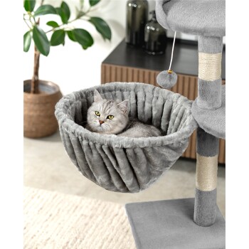 Eine flauschige graue Katze, die in einer gemütlichen, plüschigen Hängematte an einem Katzenbaum entspannt, mit einer Topfpflanze und modernen Möbeln im Hintergrund.