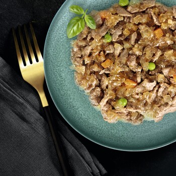 Zbliżenie na zielony talerz wypełniony jedzeniem dla zwierząt, z posiekanym mięsem, warzywami i gałązką bazylii, obok złotego widelca.