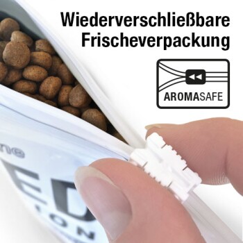Hand, die einen weißen Schieber an einem wiederverschließbaren Tierfutterbeutel mit Kroketten schließt; sichtbarer Text ‚Wiederverschließbare Frischeverpackung‘ und ‚AROMASAFE‘.