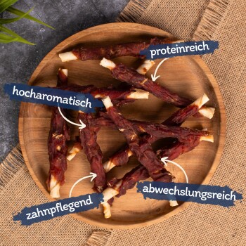 Ein Holzbrett hält mehrere Hundeleckerlis, mit Etiketten in deutscher Sprache, die anzeigen, dass sie "proteinreich", "hoch aromatisch", "Zahnpflege" und "abwechslungsreich" sind.