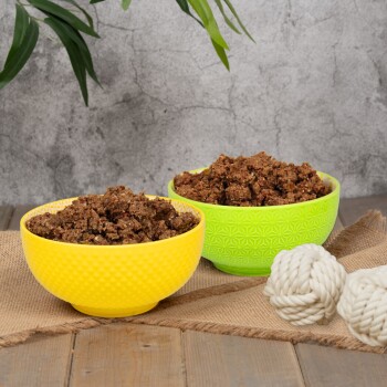 Zwei bunte Schalen, eine gelbe und eine grüne, gefüllt mit Tiernahrung, liegen auf einer Juteschicht neben weißen Seilspielzeugen.