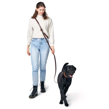 Eine Frau in einem hellen Pullover und blauen Jeans führt einen schwarzen Labrador an der Leine, lächelnd während sie gemeinsam spazieren gehen.