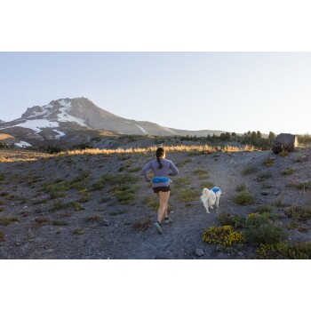 Eine Frau joggt mit einem weißen Hund an der Leine entlang eines felsigen Pfades, mit einem Berg und klarem Himmel im Hintergrund.