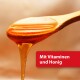 Ein Holzlöffel, der mit goldenem Honig tropft, begleitet von einer roten Box mit dem Text "Mit Vitaminen und Honig."