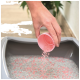 Une main verse des granulés de litière pour chats roses d'une tasse rose dans une boîte à litière grise remplie de litière grise et rose.