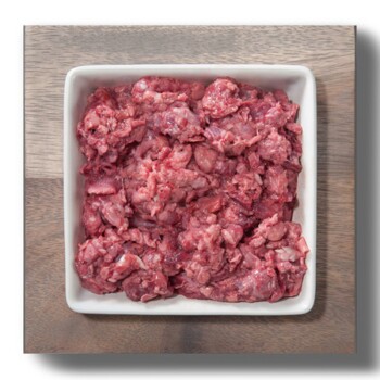 Ein quadratisches weißes Geschirr, gefüllt mit rohem, fein gehacktem rotem Fleisch, auf einer Holzoberfläche platziert.