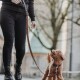 Eine Person in schwarzen Jeans und Stiefeln führt einen kleinen, flauschigen braunen Hund an der Leine und schaut auf den Hund in einer malerischen Umgebung.
