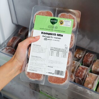 Hand hält eine gefrorene 400‑g‑Schale mit der Aufschrift ‚Komplett‑Mix Geflügel für Katzen‘, Fleischplättchen im Gefrierfach.
