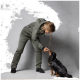 Eine Person in einem grünen Overall füttert einen kleinen schwarz-braunen Hund mit einem Leckerli, während sie vor einem grauen Hintergrund steht.