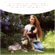 Eine Frau hält eine schwarze Katze neben einem großen Hund in einem Garten mit lila Blumen. Text: "Als Tierheilerin liebe ich die Produkte von Wolfsbacher Natur." - Lena Huber.