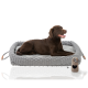 Ein brauner Labrador Retriever liegt bequem auf einem grauen gewebten Haustierbett mit einem weichen Innenraum. Ein beiges Spielzeug mit einem herzförmigen Ausschnitt liegt neben dem Bett.