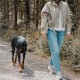 Person in beige Jacke und Jeans führt einen schwarz-braunen Dobermann an der Leine entlang eines bewaldeten Kieswegs.