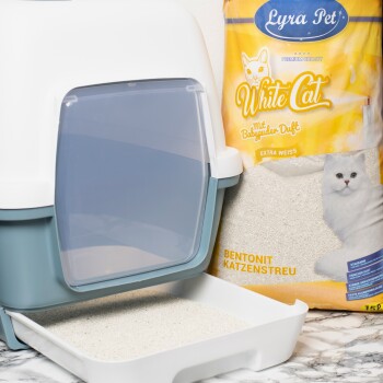 Eine Katzenstreu-Box mit einer transparenten Klappe, neben einem Sack Lyra Pet White Cat Streu, mit einem Bild einer weißen Katze und Text auf Deutsch.