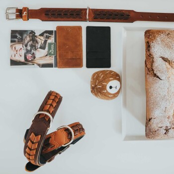 Ein stilvoller brauner Hundehalsband mit einem geflochtenen Design, zwei Lederkartenhaltern (einer braun, einer schwarz), einem Plüschspielzeug und einem Kuchen auf einem weißen Teller.