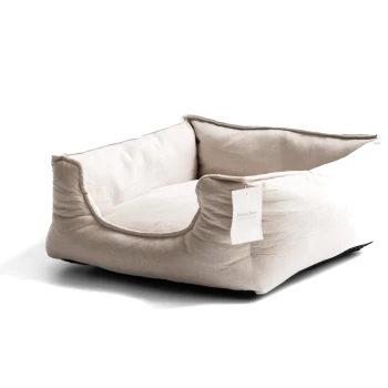 Schlafplatz Körbchen Katzenbett Linen Ein gemütliches, beiges Haustierbett mit erhöhten Kanten und einem weichen Kissen, mit einem kleinen Etikett, das "Muta & Mela" liest.