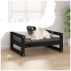 Ein entspannter Mops, der auf einem stilvollen schwarzen Haustierbett mit einem grauen Kissen liegt, neben einem grünen Tennisball und einer Topfpflanze.