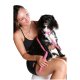 Eine Frau in einem schwarzen Tanktop und Shorts hält einen kleinen schwarz-weißen Hund mit einem rosa Geschirr und einer Leine, der auf einem weißen Hocker sitzt.