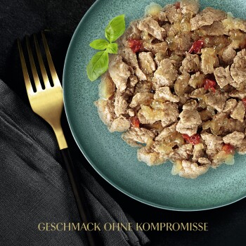 Ein Teller mit Haustierfutter, das zerrissenes Fleisch mit Gemüse vermischt zeigt, garniert mit einem Basilikumblatt, mit dem Text "GESCHMACK OHNE KOMPROMISSE" darunter.