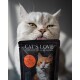 Puszysty szary kot liże wargi, trzymając paczkę karmy dla kotów "CAT'S LOVE", z zdjęciem pomarańczowego i białego kota.
