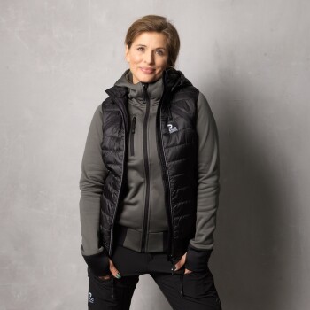 Eine Person trägt eine schwarze, gepolsterte Weste über einem grauen Reißverschluss-Hoodie, mit Händen in den Taschen, vor einem neutralen Hintergrund.