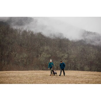 Eine Frau in einer türkisfarbenen Jacke und ein Mann in einer blauen Jacke führen einen schwarzen Hund auf einer grasbewachsenen Fläche mit einem nebligen Wald im Hintergrund.