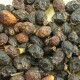 Nahaufnahme verschiedener Trockenfrüchte, überwiegend dunkelbraun und schwarz, mit einigen rötlichen Farbtönen, verstreut auf einer hellen Holzoberfläche.