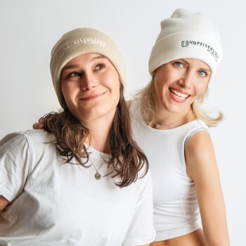 Zwei Frauen tragen beigen Mützen und weiße Oberteile, lächeln und posieren eng vor einem schlichten weißen Hintergrund.