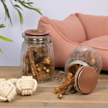 Eine schwarze Schüssel mit getrockneten Hundekauartikeln auf einer grauen Oberfläche, umgeben von verstreuten Kauartikeln, einem geflochtenen Korb und einem weißen Seilspielzeug.