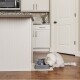 Un chat blanc duveteux boit dans une fontaine à eau pour animaux grise sur un tapis beige dans une cuisine moderne avec des armoires claires et un plan de travail en granit.