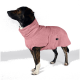 Ein brauner Hund, der einen weichen rosa Haustier-Hoodie trägt, steht an einem sandigen Strand, umgeben von hohem Gras und einem verschwommenen Hintergrund.