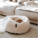 Ein kleiner Hund, der sich in einem flauschigen, runden Hundebett auf einem hellen Teppich zusammengekuschelt hat, mit einem gemütlichen Sofa im Hintergrund.