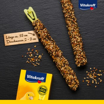 Deux bâtonnets de graines pour oiseaux, d'environ 15 cm de long et 2-3 cm de diamètre, avec un clip jaune, à côté d'une boîte Vitakraft jaune.