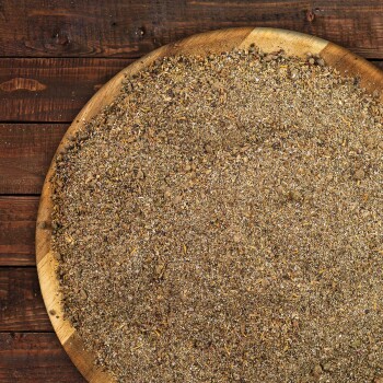 Eine Holzschüssel gefüllt mit einer groben, bräunlich-grauen Mischung aus Kräutern und Gewürzen, vor einem rustikalen Holz-Hintergrund.