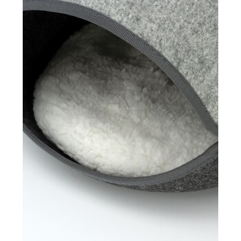 Ein gemütliches, graues Haustierbett in Form einer Kuppel steht auf einem Holzboden, mit einer teilweise sichtbaren Katze darin. Im Hintergrund ist eine Topfpflanze.