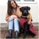 Eine Frau in einem beigen Mantel sitzt auf einer Steinmauer neben einem schwarzen Hund, der eine kastanienbraune Jacke mit einer gefütterten Kapuze trägt.