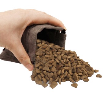Hand gießt braunes, dreieckiges Trockenfutter aus einem kleinen grauen Stoffbeutel.