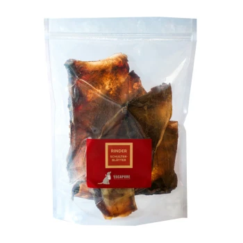 Eine durchsichtige Plastiktüte mit Hundeleckerlis aus Rinderschulterblättern, beschriftet mit "RINDER SCHULTER-BLÄTTER" auf rotem Hintergrund.