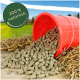 Ein gekippter roter Eimer, der grüne Tierfutterpellets auf Holz verschüttet, mit einem Feld und blauem Himmel im Hintergrund. Text sagt "100 % natürlich."