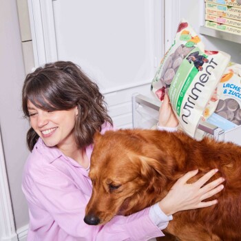 Eine Person in einem pinken Shirt hält eine Tüte "Nutriment" Hundefutter, während sie einen Golden Retriever in einem Tiergeschäft umarmt.